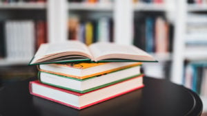 A photo of several books stacked on top of a small table, with more books on shelves in the background.